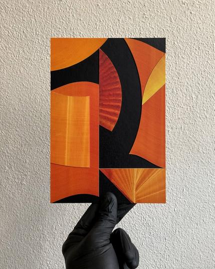 A gloved hand holds a collage-art on a wall. The collage shows orange and yellow geometric paper-cutout shapes on a black background.