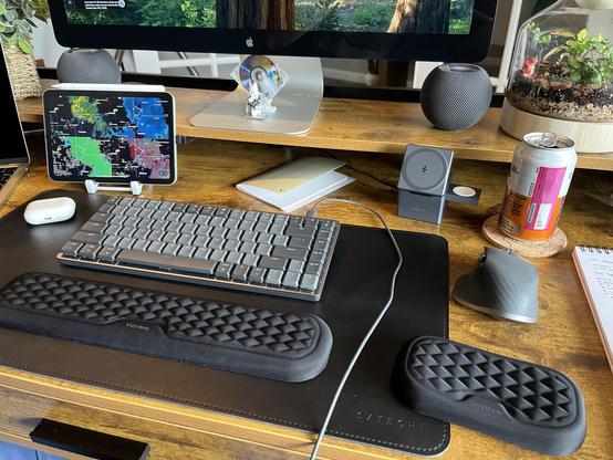 A photo of a desktop of red-brown wood grain with a black leather mat covering most of the desk with two separate black wrist rests for a keyboard and mouse. The prominent mechanical keyboard chonky mouse are in black and gray tones. There is an iPad behind the keyboard to the left showing the weather radar app Radar Scope. The bottom of a Thunderbolt Display is at the top of the frame, flanked by two black iPod minis. A terrarium of blacks, browns, and greens in on the top-right corner with a can of Mai Tai directly in front of it.

Oh and white lego Batman, also known as bachelor batman, is sitting in front of a silver and clear minidisc on top of the stand of the Thunderbolt Display.