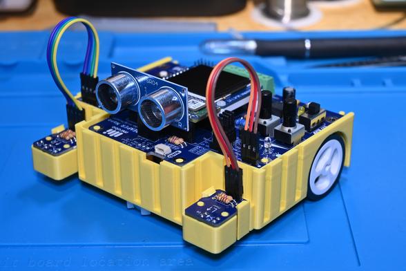 A yellow, 3d-printed BEAPER Bot robot controlled by an Arduino Nano ESP32 mounted on a blue BEAPER Nano PCB (hidden behind the SONAR distance sensor module). Two optical floor sensors are connected by extension cables at the front of the robot (left) and a white drive wheel is visible at the back.