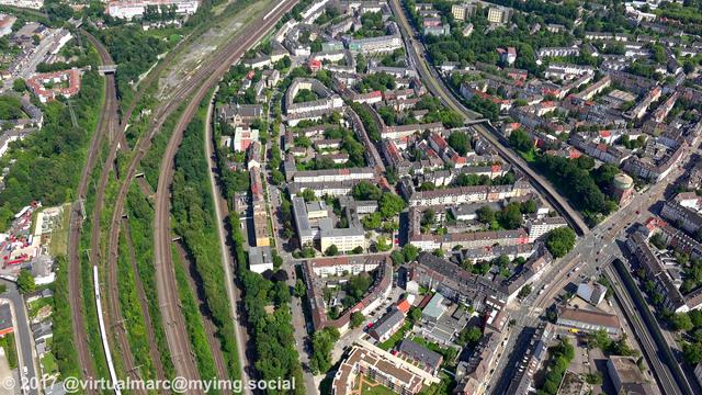 An aerial photo of the center of essen with houses and workplaces sourrounded by trees. The main freeway (the A40, ruhr express way, Ruhrschnellweg) can be seen on the right. Multiple Train tracks can be seen on the left.