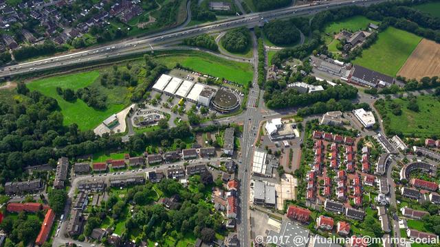 An aerial view of Recklinghausen. A freeway can be seen at the top, multiple living quaters sourrounded by trees and grass are at the bottom. There are some company buildings next to the freeway.