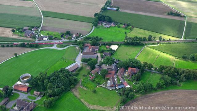 An aerial view of the countryside of Recklinghausen with some houses in between sourrounded by trees. A railway track is going through the image. A red train is driving on the track.