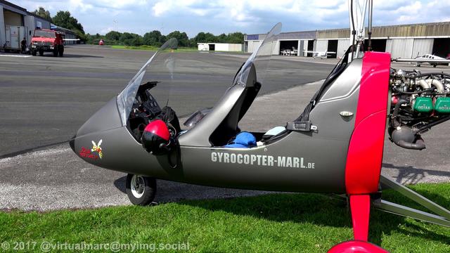 A photo of the gyrocopter from the side, parking on a grass area near the hangars. There are some sports planes in front of the hangars and a helicopter in the back. A fire truck is standing next to a hangar.