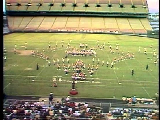 A drum and bugle corps performance is taking place on a football field inside a stadium. The corps is arranged in various formations, with a crowd of spectators seated in the stands. The field features some markings typical of a sports arena.