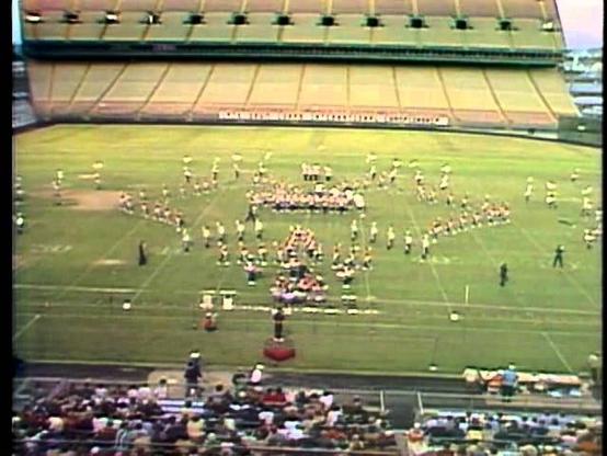 A drum and bugle corps performance is taking place on a football field inside a stadium. The corps is arranged in various formations, with a crowd of spectators seated in the stands. The field features some markings typical of a sports arena.
