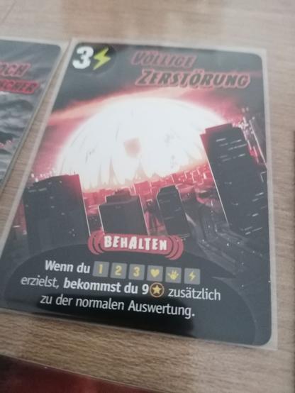 Eine Spielkarte mit dem Namen Völlige Zerstörung. Die Illustration zeigt eine Großstadt in deren Mitte eine gewlatige Explosion zu sehen ist. Der Effekt im Spiel sind der Erhalt von 9 Punkten. Dies führt in vielen Fällen zum Spielsieg, wofür 20 Pkt. nötig sind.