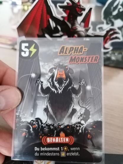 Zusehen ist die Spielkarte mit dem Namen Alpha-Monster. Es zeigt eine uberdimensional große Ratte, die scheinbar laut schreiend auf ihren Hinterbeinen steht. Vor ihr laufen unzählige kleinerer Ratten. Der Effekt der Karte bringt einen Extrapunkt, wenn ein Schaden gewürfelt wird.