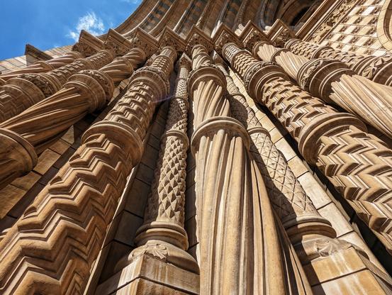 A low-angle shot looks up at the ornate facade of a grand, tan-coloured stone building. Multiple columns, each uniquely carved with intricate patterns such as zigzags, spirals, and textured surfaces, rise upwards towards an arched structure at the top. Sunlight catches the details of the stonework, creating highlights and shadows that accentuate the depth and texture of the carvings. A patch of bright blue sky is visible in the upper left corner, contrasting with the warm tones of the building. The architectural style appears historical and elaborate.