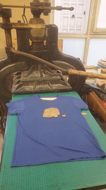 T-shirt posé à plat sur le plateau d'une énorme presse, linogravures posées face contre tissu