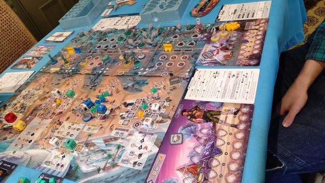 Perseverance, Castaway chronicles, Episode one mid-game. The board shows a beach with various worker-placement (dice) locations which can be used to gather resources, send out soldies on patrol, train more soldiers. After the beach there are defensive positions for soldiers and walls, and attacking dinosaur models