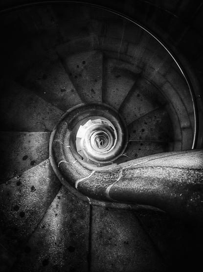 A high-angle, black and white shot of a stone spiral staircase winding downwards towards a bright light at the center. A metal handrail curves along the outer edge of the steps.