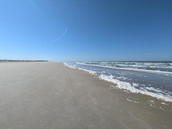 Foto von einem leeren Strand, von rechts kommt das Wasser, links am Horizont sind Dünen.