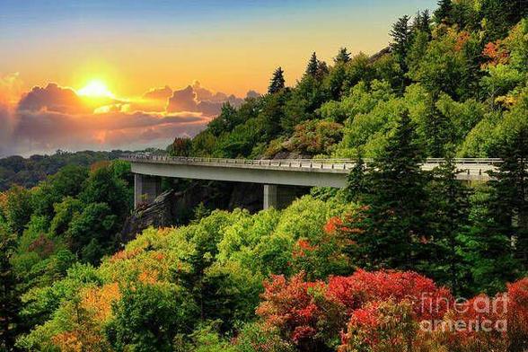 Scenic autumn view of the Blue Ridge Parkway's Linn Cove Viaduct near Grandfather Mountain, North Carolina, at sunset. The curving bridge stretches over a vibrant landscape of orange, yellow, and red fall foliage against the backdrop of the Blue Ridge Mountains. The warm, golden light of the setting sun enhances the rich autumn colors, creating a serene and breathtaking mountain vista.