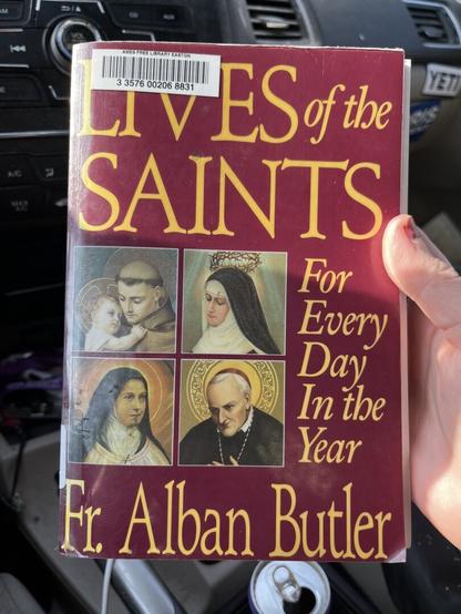 a red softcover book, featuring icons of a few various saints. in gold script, the title is “LIVES OF THE SAINTS: For Every Day In the Year”, by father alban butler.