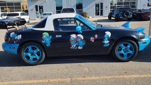 A small sports car perhaps from 1980's painted black with bright blue highlights. It is covered with nude female smirf cartoon characters, and excited male smirfs. it is in a parking lot in front if a building. It is a sunny day.