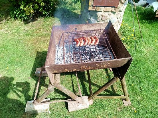 BBQ in action. The two stands, now leveled with some old bricks have the trough standing on them (it fits in the gaps between the joined pipes). Inside are embers from a wood fire and a large grille suspending a smaller one. On that, six thick sausages, nicely brown already.