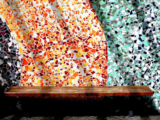 This image shows a beautiful mosaic wall with a wooden bench in front of it. The mosaic features two distinct color schemes - the left side consists of vibrant orange, red, and yellow tiles, while the right side transitions to cool mint green, turquoise, and white pieces. The contrast between the warm and cool colors creates a striking visual effect.
The wooden bench appears to be made of rustic, weathered wood with a simple design, mounted against the colorful backdrop. The bench sits on what looks like a stone or paved surface at the bottom of the frame.