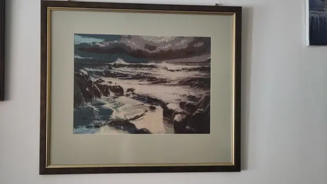 A framed cross stitched artwork of a seaside scene with heavy storm and a wild sea. The original is Edward Barton's "sanctuary".