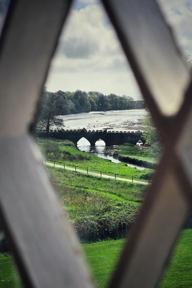 A stone arch bridge spans a small river, viewed through a diamond-shaped window pane. Green fields and trees line the riverbanks under a cloudy sky.
