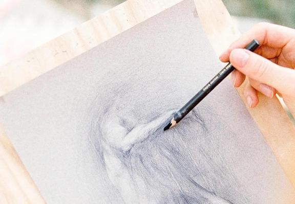 The image shows a close-up of a hand holding a black pencil, drawing on a piece of paper mounted on a wooden easel. The drawing depicts a figure with a draped garment, focusing on the shoulder and upper arm area. The pencil is being used to add shading and texture, particularly around the folds of the fabric. The paper is light gray, and the background is blurred, suggesting an outdoor setting. The hand is steady, indicating a careful and deliberate drawing process. The pencil appears to be a standard graphite pencil, with a sharpened tip, and the drawing shows a range of tones from light to dark, creating a sense of depth and form.

Provided by @altbot, generated privately and locally using Ovis2-8B