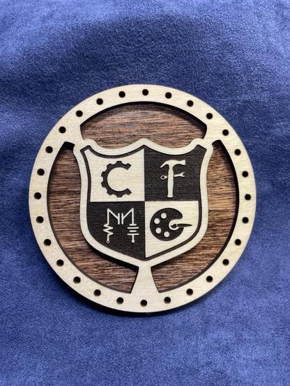 Picture of a round laser engraved and cut disc.  Made from 2 layers with the back layer stained dark brown. The top layer is clear coated natural wood with the shield shaped maker space logo engraved in the center. The area around the shield is cut out except for 3 attachment points to the rim. All around the rim are 2mm holes spaced about 10 mm apart.