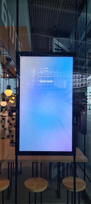 A large rectangular, portrait oriented, black bordered display unit hanging between two, vertical, black rails in the window of a lunchroom. 

In the top third of the display a small white circle with a picture of a usb c port above the text:

Display Port
No cable connected
(1) Check that the cable is plugged in and the settings of the source device are correct.
(2) Press Source below to select a connected source

Below the text a rectangular, white, landscape oriented button with the word source in it.

All this centered in the middle against a full display, tie dye like, pastel blue and purple background.