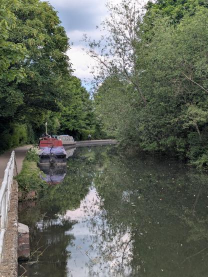 The image depicts a serene canal scene. In the foreground, a narrowboat with a red cabin is moored on the left side of the canal, partially covered with a purple tarp. The water is calm, reflecting the surrounding greenery and the overcast sky. A white metal railing runs along the canal's edge, separating the path from the water. On the path, a person is seen walking away from the camera, adding a sense of scale to the scene. The canal is flanked by dense trees and bushes, creating a lush, green environment. The sky is partly cloudy, with patches of blue visible through the clouds. The overall atmosphere is peaceful and tranquil, with the natural elements harmoniously blending with the man-made canal.