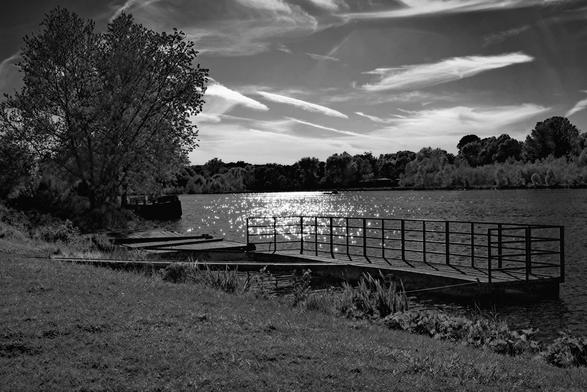 The image depicts a black and white scene of a lakeside or riverside with a wooden dock extending into the water. There's a grassy foreground leading to the dock, which has a railing. Reflective sunlight is glistening on the water's surface. Trees line the opposite shore, and the sky is filled with wispy clouds.