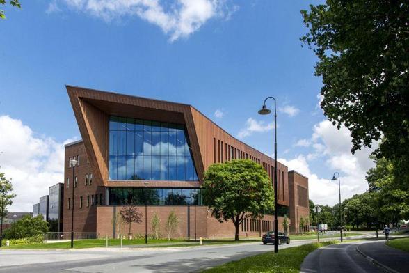 Photograph of a large building with a glass front facing the camera. The building is a light brown colour, a mixture of brick and metal cladding. There is a blue sky overhead