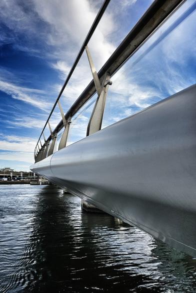 A low-angle, wide shot shows a contemporary grey bridge extending over dark, rippled water in Belfast under a bright blue sky with wispy white clouds. The bridge features sleek, curved sides and metal railings. In the distance, across the water, are indistinct buildings and structures typical of the Belfast waterfront.