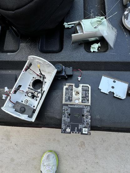 An assortment of electronic components, including a disassembled device casing, a circuit board, and some loose parts, are scattered on a surface. A shoe partially visible at the bottom adds context.