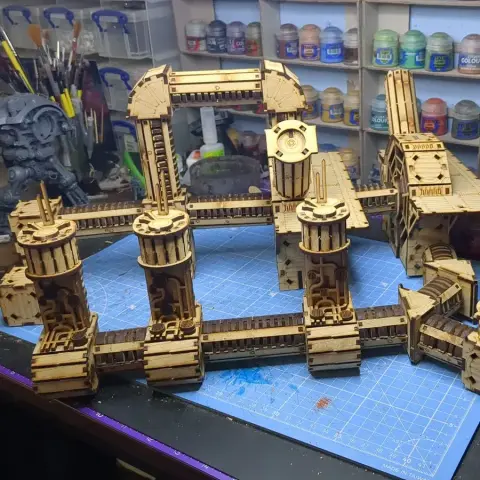 A detailed assembly of the TT Combat Sector 4 Plasma Relay is displayed, featuring intricate wooden structures with multiple towers and connecting pathways. The design includes cylindrical towers with circular tops and various platforms, all set against a blue cutting mat. In the background, jars of paint and tools are visible, indicating a workspace dedicated to tabletop gaming and model building.