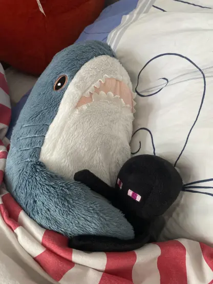 A cozy BLÅHAJ shark plushie tucked into bed, cuddling with a small black Minecraft Enderman plush toy. The IKEA shark's blue-gray fuzzy body, white underbelly, and signature pink open mouth with felt teeth are prominently displayed as it appears to be protectively embracing its blocky companion. The Enderman with its characteristic purple eyes is nestled against the shark's side. Both plushies are resting on white bedding with blue line details and a red and white striped blanket or pillow. A sweet bedtime scene showing that even apex predators of the deep need cuddle buddies for a good night's sleep!