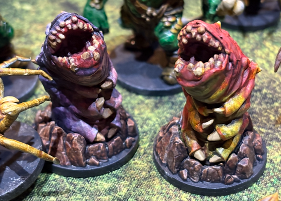 Photo of two miniatures of subterranean monster worms coming up out of the ground. They have big maws of jagged teeth and six little talons arms. One is purple and one is red.