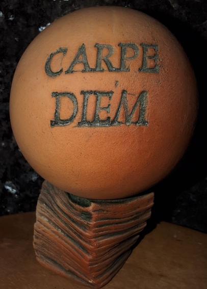 eine Tonkugel mit einer angenehm hellbraunen bis rotbraunen Färbung.
Auf der Tonkugel steht in grünmetallisch wirkenden Großbuchstaben "CARPE DIEM"
Die Tonkugel liegt auf einem eigens für sie hergestellten Tonsockel, der ca. 50% kleiner ist als der Durchmesser der Kugel. Der Sockel sieht aus wie aus mehreren Lagen dickeren, übereinandergelegten, quadratischen Papierseiten, ebenfalls in rotbraunen und grünen Streifen