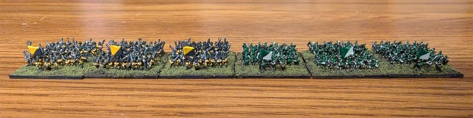 Photo of twelve bases of 3 mm fantasy miniatures from Microworld Games arranged in a 6x2 line. On the left are Doppelsöldner units in yellow and black; on the right are Swordsmen in shining armor with green shields and plumes, with green and white standards.