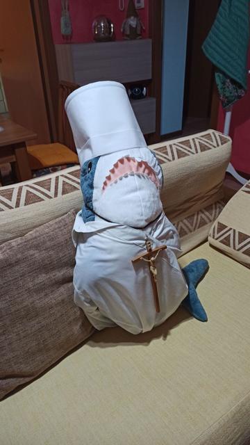 Holy BLÅHAJ! An IKEA shark plushie dressed as a Catholic priest or pope, complete with white robes, a tall cylindrical hat resembling a mitre, and a wooden crucifix hanging around its neck. The blue-gray shark with its characteristic white underbelly and pink mouth is seated on a beige carpet next to a geometric patterned couch. Its fin peeks out from beneath the religious attire, maintaining its shark identity despite the holy transformation. In the background, a living room with furniture and decorative items is visible. This shark has clearly taken its vocation seriously, ready to deliver a sermon to its plush congregation.