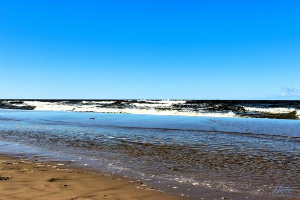 Im Vordergrund seht ihr ein Stückchen vom Ostseestrand, danach seichtes Wasser, nach der ersten Sandbank im Wasser seht ihr große Wellen. Spaß für Wasserratten. Ansonsten geht das dunkelblaue Wasser in einen hellblauen Himmel über.