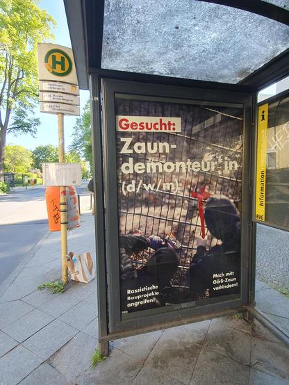 Ein Buswartehäuschen von der Seite, im strahlenden Sonnenschein.  Auf der Fläche, auf der sonst Werbeplakate sind, ist ein Plakat im Stil einer Stellenanzeige. 
Text: "Gesucht: Zaun-Demonteur*in (d/w/m). 
Unten steht klein: Rassistische Bauprojekte angreifen! Mach mit: Görli- Zaun verhindern!.
Das Foto  zeigt eine rote Zange, die einen Maschendrahtzaun durchschneidet.