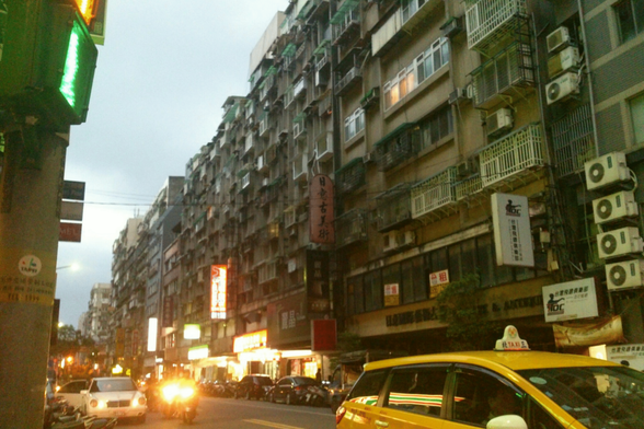 Street scene that consists of a narrow road with highrises on all sides, cluttered facades of windows and air conditioning units. Cars, yellow taxis and traffic in the foreground, a narrow stretch of pale sepia evening sky above it. The high buildings make you feel small. Overall atmosphere is a dense and busy city, with no trees or  much space to breathe. Not a place to stay but to pass through to elsewhere. Colours are mostly yellow-ish from the setting sun and car headlights.