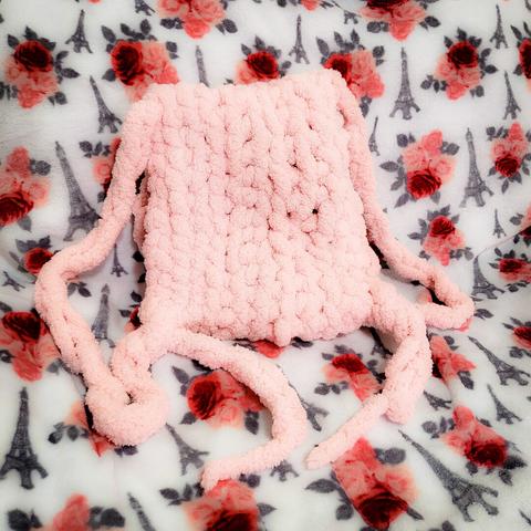 - white blanket with patterns: grey Eiffel Towers, red/pink roses with grey leaves
- back-view of a soft pink fluffy crocheted pouch
- two long strands on both sides are tied at the edges of the pouch, to represent a backpack style