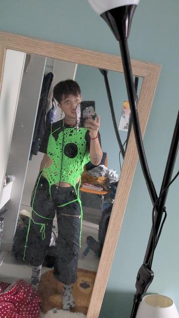 Une personne debout tient son téléphone et est encadré.e par un miroir. Iel porte un haut en crochet vert fluo avec un trou au milieu du torse, et des cordons noués autour des jambes