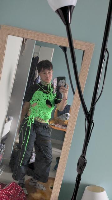 Une personne debout tient son téléphone et est encadré.e par un miroir. Iel porte un haut en crochet avec des trous au niveau du torse et des cordons noués autour des hanches et du cou