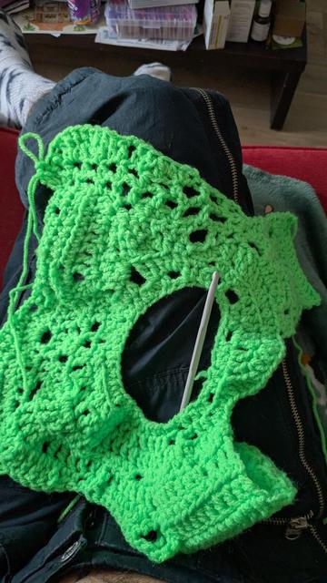 On voit une pièce de crochet vert fluo de haut, posée sur des jambes croisées. Un crochet métallique est posé au milieu ce qui laisse penser qu'elle est en cours de fabrication