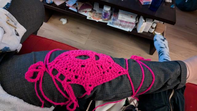 On voit une jambe étendue vêtue d'un pantalon et , par dessus le pantalon une pièce en crochet rose fluo avec une forme de losange autour du genou et des cordons noués autour de la cuisse et du mollet