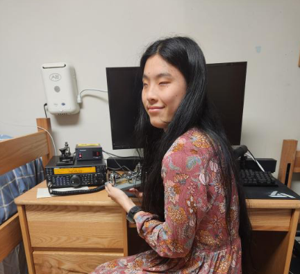 A young, blind, Asian woman sitting in front of a wooden desk with a TS-590S radio on it, holding a Vibroplex semiautomatic key in her hands.