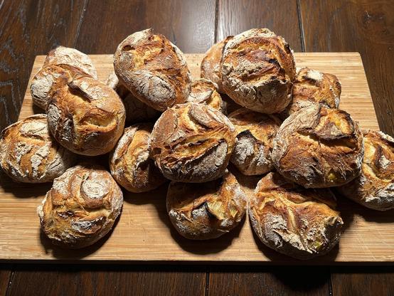 Auf einem dunklen Holztisch liegt ein Holzbrett. Darauf liegen zahlreiche frisch gebackene Brötchen mit rustikalem Ausbund.