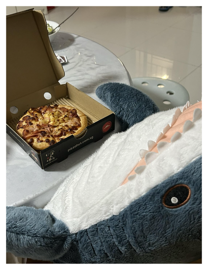 BLÅHAJ's pizza night! An IKEA shark plushie lying on what appears to be a glass table, eyeing a small personal pizza from Pizza Hut Singapore (visible from the pizzahut.com.sg on the box). The blue-gray shark with its distinctive white underbelly, pink mouth, and felt teeth is positioned with its embroidered black eye clearly focused on the cheesy prize. The open pizza box reveals what looks like a ham or bacon topped pizza with melted cheese. The shark's mouth is perfectly positioned as if ready to dive in for a bite.
