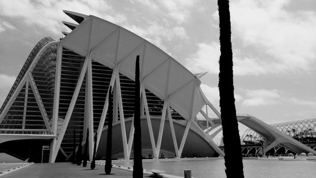 A black and white photograph shows a modern, angular building with a prominent white skeletal structure against a cloudy sky. Tall, slender dark trees stand in front of the building, partially obscuring the view. A body of water reflects a portion of the building and the sky. A paved walkway leads towards the structure, and indistinct figures of people can be seen near the base of the building.