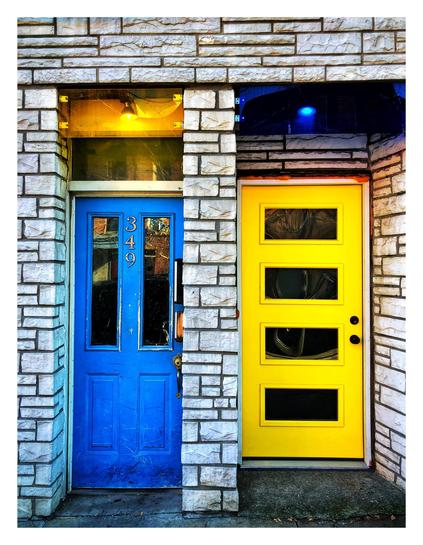 A blue door and a yellow door, set into a facade of differently sized stone bricks forming irregular grid patterns. The doors complement each other, as the blue door features narrow vertical glass panels while the yellow door has horizontal ones. Both also have transom windows with lights shining inside, a yellow light over the blue door and a blue light over the yellow door. The number “349” appears vertically on the middle top section of the blue door.